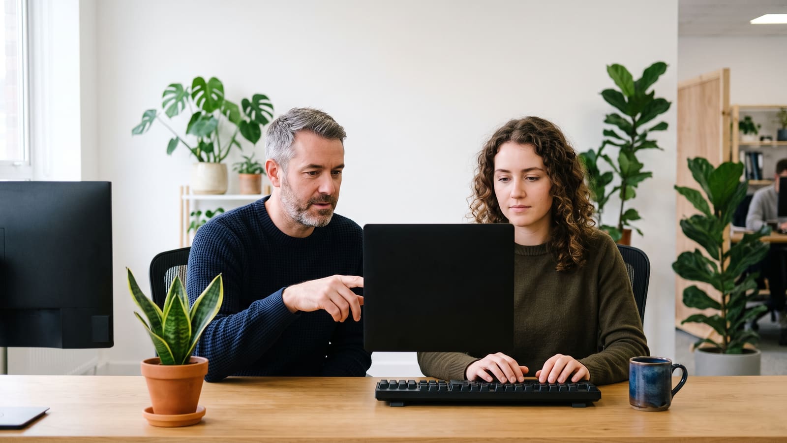 Two developers pair-programme at one desk, one pointing at the monitor
