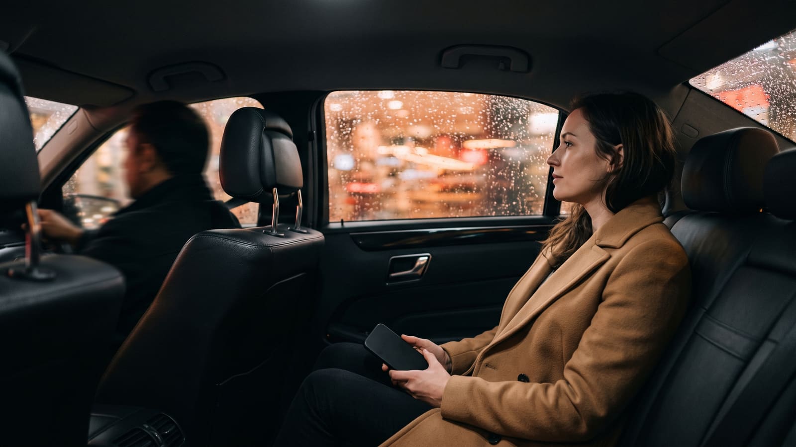 A woman in the back seat of a private hire car looks out at city lights on a rainy evening